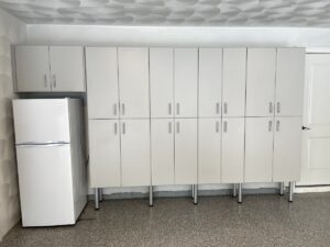 A row of modern white custom garage cabinets with a small refrigerator to the left, set against a neutral wall.