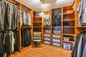 A neatly organized walk-in closet with wooden shelves and racks, displaying a variety of clothes, shoes, and storage boxes. A chair is placed in the center of the closet.
