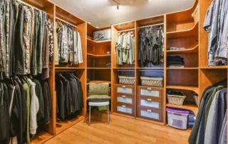 A neatly organized walk-in closet with wooden shelves and racks, displaying a variety of clothes, shoes, and storage boxes. A chair is placed in the center of the closet.