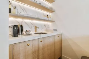 Compact kitchen area with hidden pantry, butler's pantry, wooden cabinets, a white countertop, and a small sink. Marble backsplash features open shelves with various kitchen items. A coffee machine and decor items are placed on the counter.