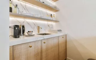 Compact kitchen area with hidden pantry, butler's pantry, wooden cabinets, a white countertop, and a small sink. Marble backsplash features open shelves with various kitchen items. A coffee machine and decor items are placed on the counter.