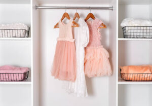 Three children's dresses—two pink and one white—hanging on wooden hangers in a white closet, with wire baskets containing folded clothes on either side.