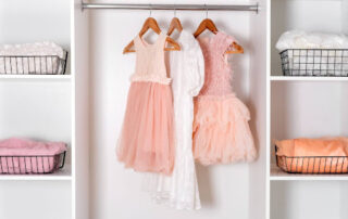 Three children's dresses—two pink and one white—hanging on wooden hangers in a white closet, with wire baskets containing folded clothes on either side.