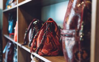 Assorted handbags displayed on wooden shelves, featuring various colors and textures, including a prominent red bag with a distinctive chain strap.
