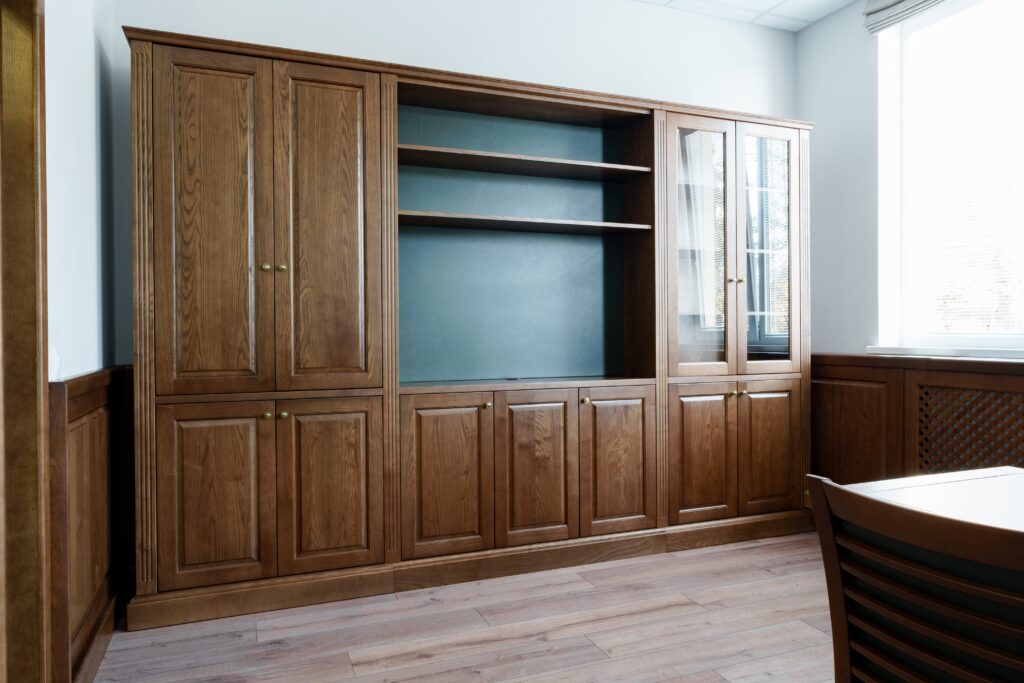Wooden office cabinet with closed doors, open shelves, and a glass-paneled section accentuating its design. Adjacent to the cabinet is a wooden table and chair on a light wood floor, creating an inviting workspace reminiscent of custom entertainment centers.