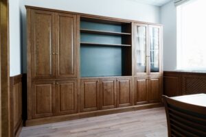 Wooden office cabinet with closed doors, open shelves, and a glass-paneled section accentuating its design. Adjacent to the cabinet is a wooden table and chair on a light wood floor, creating an inviting workspace reminiscent of custom entertainment centers.