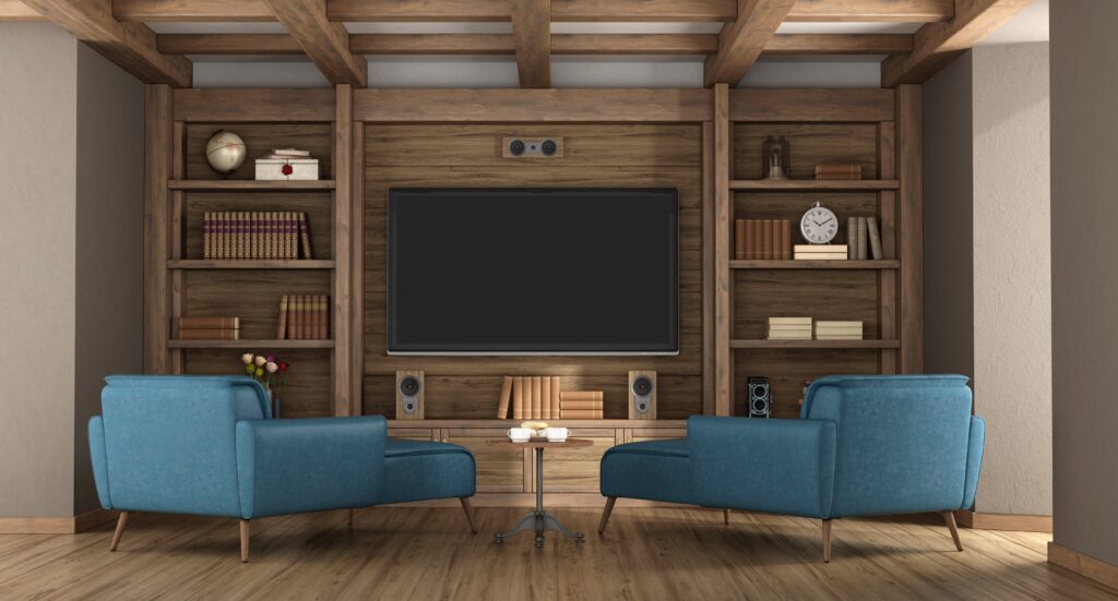 A modern living room with a large flat-screen TV, two blue armchairs, wooden shelves with books, a clock, and decorative items, all set on a wooden floor with exposed beams on the ceiling.