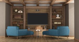 A modern living room with a large flat-screen TV, two blue armchairs, wooden shelves with books, a clock, and decorative items, all set on a wooden floor with exposed beams on the ceiling.
