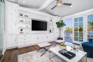 Modern living room with white built-in shelving, a large TV, and glass doors leading to a balcony. Features a ceiling fan, potted plant, and a white coffee table with books and a moss centerpiece.