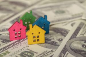 Four small wooden house models in red, green, blue, and yellow sit atop scattered hundred-dollar bills, echoing how custom closets can increase home value.