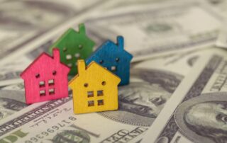 Four small wooden house models in pink, green, blue, and yellow are placed on a background of US dollar bills, symbolizing how thoughtful additions like custom closets can increase home value.