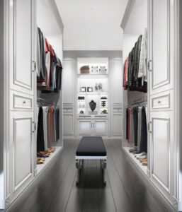 A spacious walk-in closet with custom white cabinetry, clothes on hangers, folded garments, shoes on shelves, and a bench in the center showcasing how to design a custom walk in closet