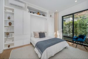 A modern bedroom with a neatly made bed, built-in white shelving, decorative items, a blue chair, large windows, and a view of greenery outside.