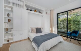 A modern bedroom with a neatly made bed, built-in white shelving, decorative items, a blue chair, large windows, and a view of greenery outside.