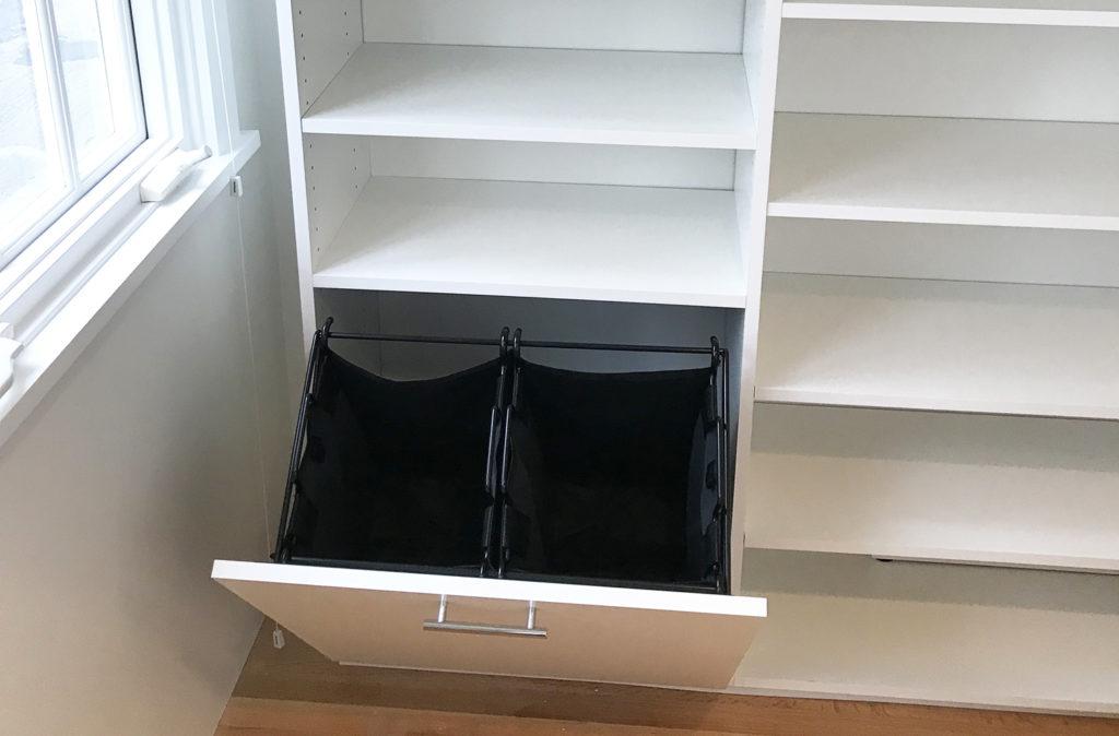 Open white cabinet with two black fabric laundry hampers inside, installed beneath shelves next to a window.