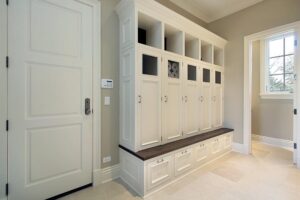 Built-in white mudroom locker storage with individual cubbies, upper open shelves, closed cabinets, and a dark wooden bench in a room with beige walls and tile flooring.