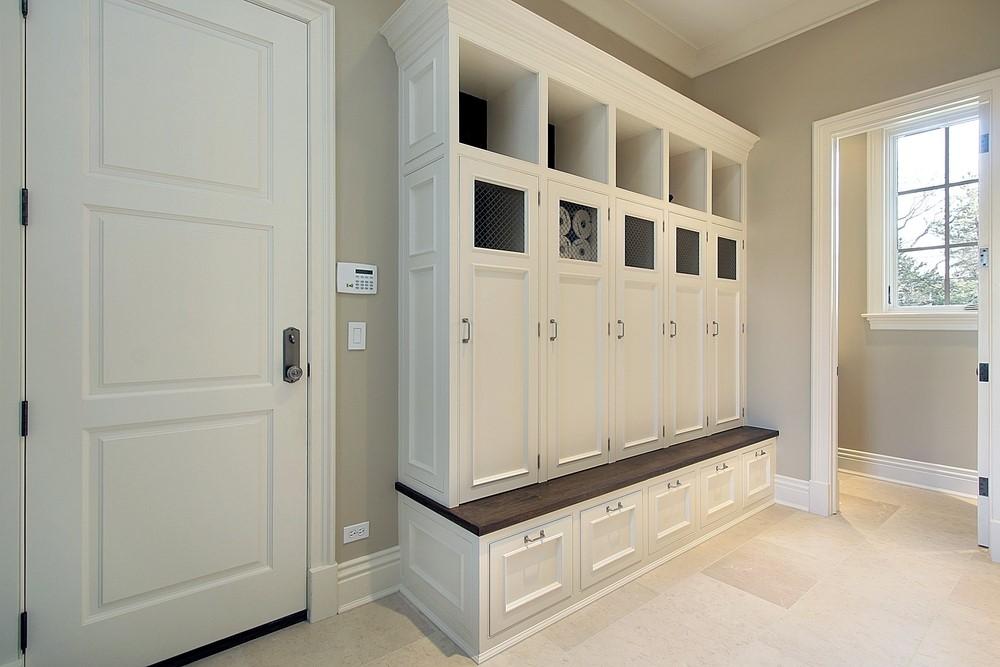 Built-in white mudroom locker storage with individual cubbies, upper open shelves, closed cabinets, and a dark wooden bench in a room with beige walls and tile flooring.