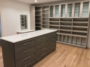 A spacious walk-in closet with built-in shelves, glass-front cabinets, and a large island with drawers on a light wood floor.