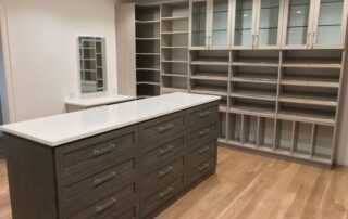 A spacious walk-in closet with built-in shelves, glass-front cabinets, and a large island with drawers on a light wood floor.