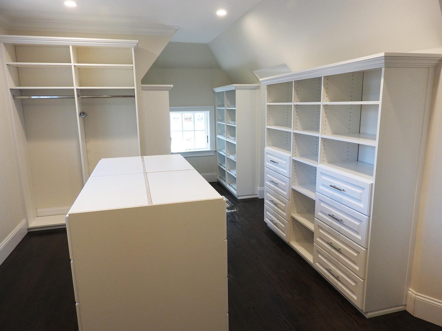 A spacious walk-in closet with built-in white shelves, drawers, and open storage spaces, dark wood floor, and a window providing natural light.