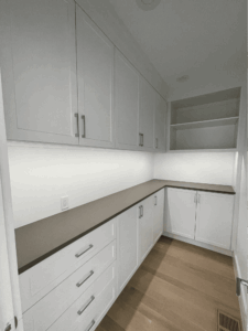 A modern kitchen pantry with white cabinets, drawers, a beige countertop, open shelves, and light wood flooring.