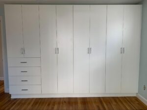A white built-in wardrobe with five doors and four drawers, featuring silver handles, stands against a light-colored wall on a wooden floor.