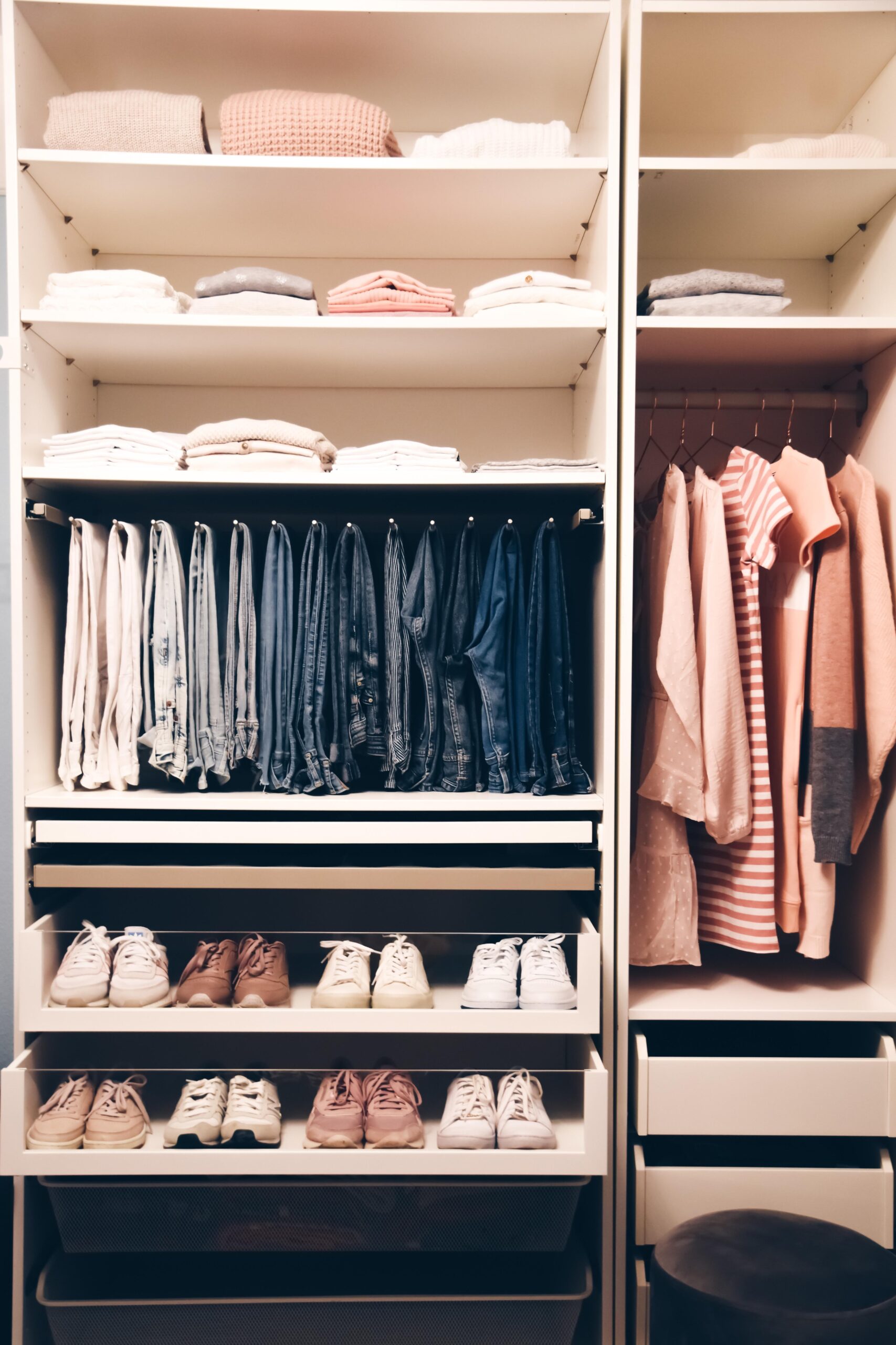 A neatly organized reach-in closet with folded sweaters and shirts on shelves, jeans hung in a row, shirts and jackets on hangers, and several pairs of shoes on pull-out racks.