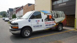 A white Boston Closet Co. van with company branding and contact information is parked outside a commercial building, ready to deliver expert reach-in closet solutions.
