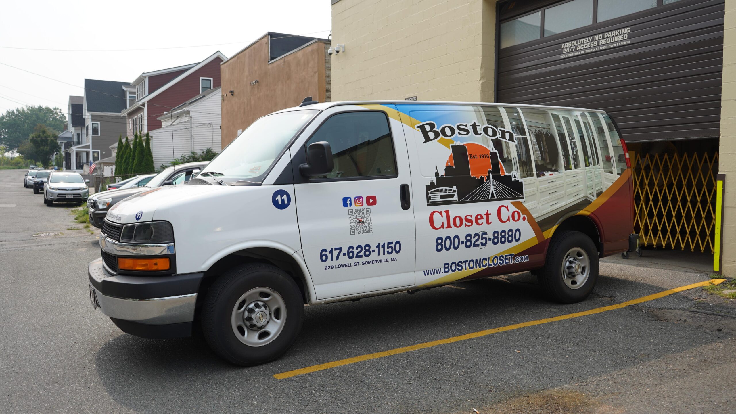A white Boston Closet Co. van with company branding and contact information is parked outside a commercial building, ready to deliver expert reach-in closet solutions.