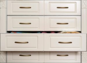 A set of six white cabinet drawers with brass handles, perfect for a reach-in closet; three are slightly open, revealing various colorful items inside.