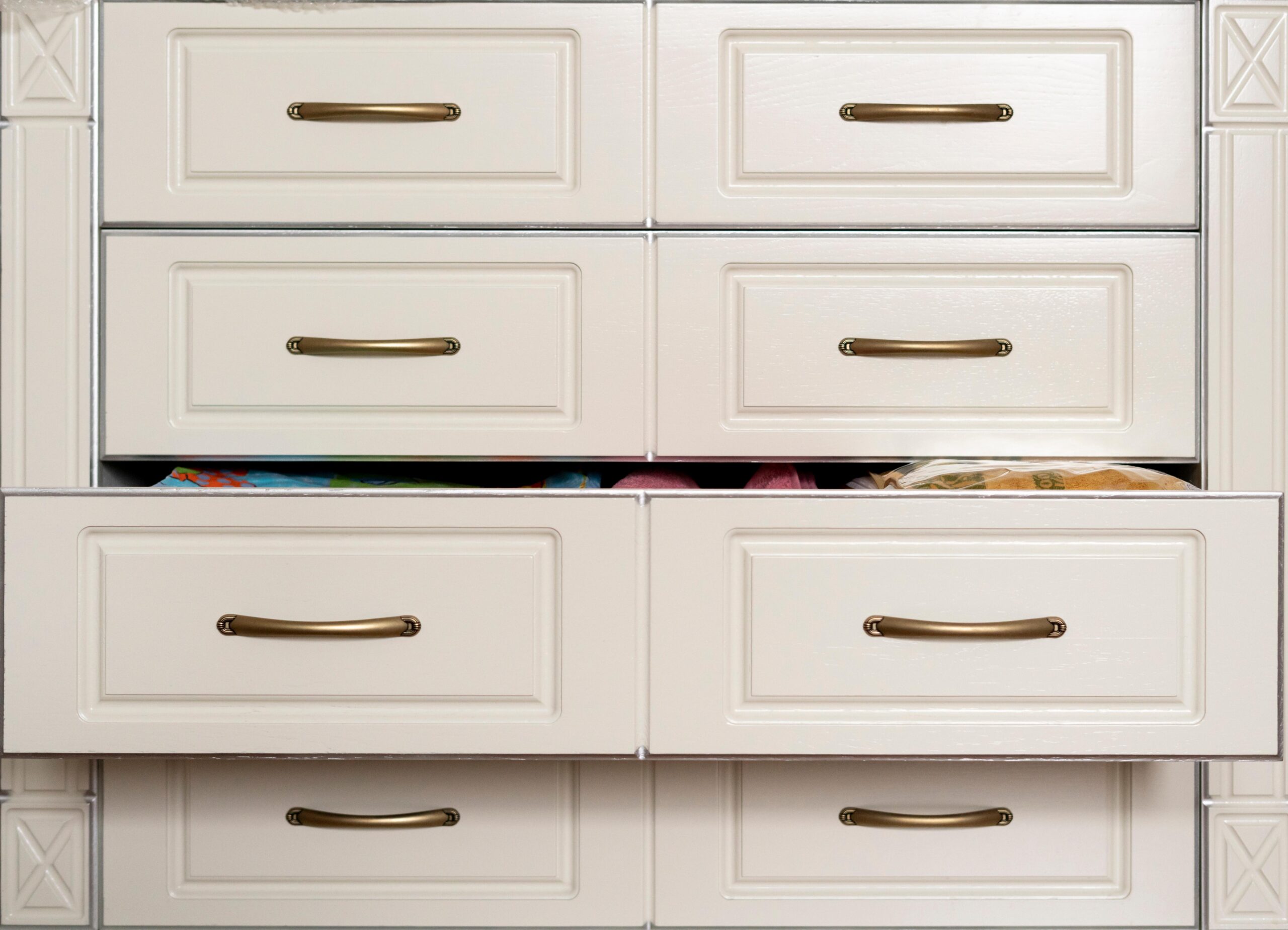 A set of six white cabinet drawers with brass handles, perfect for a reach-in closet; three are slightly open, revealing various colorful items inside.