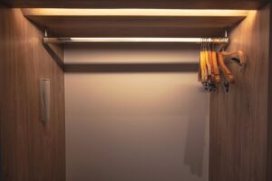 Empty wooden reach-in closet with a horizontal metal hanging rod, several wooden hangers on the right side, and overhead lighting illuminating the interior.