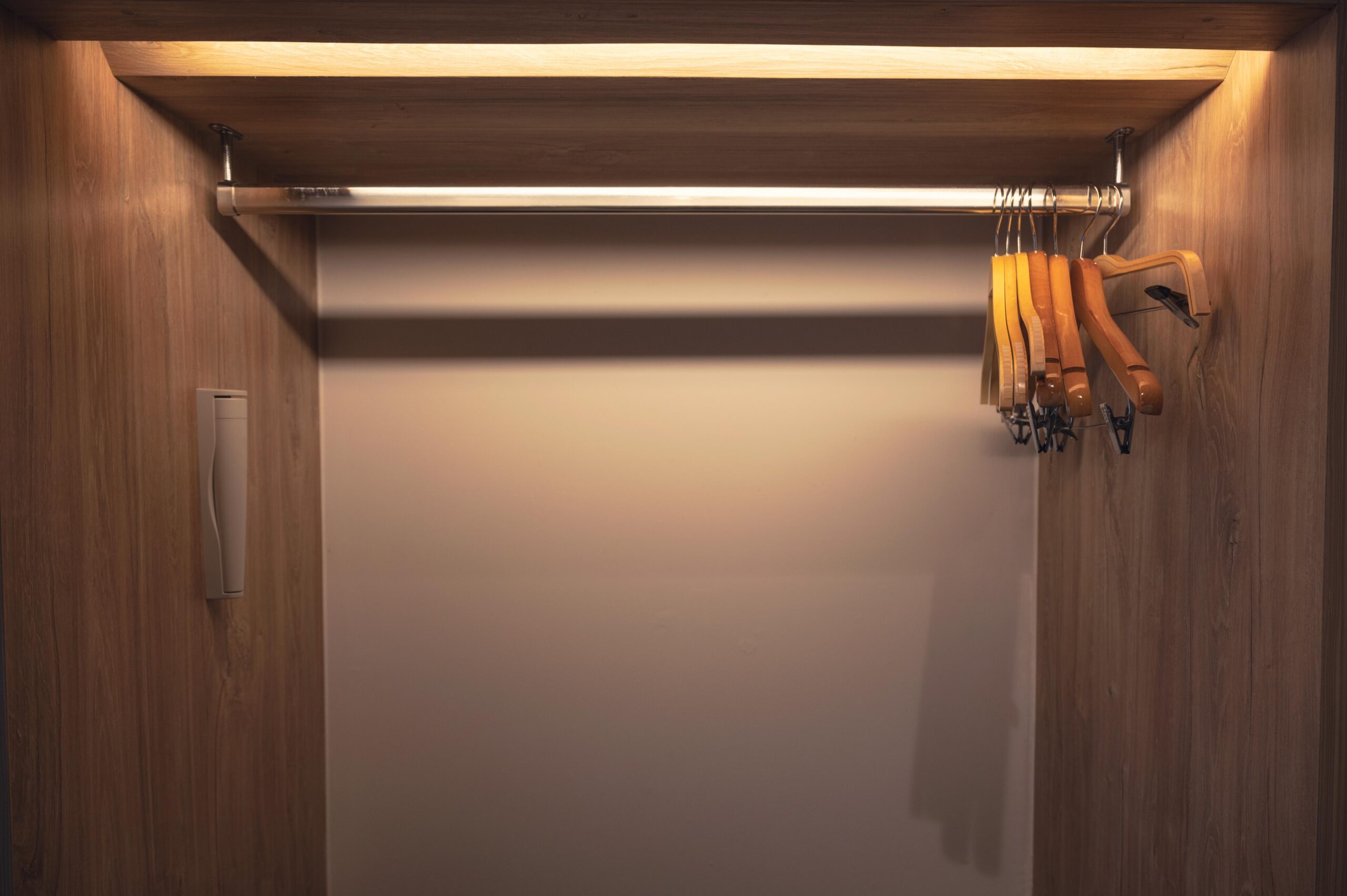 Empty wooden reach-in closet with a horizontal metal hanging rod, several wooden hangers on the right side, and overhead lighting illuminating the interior.