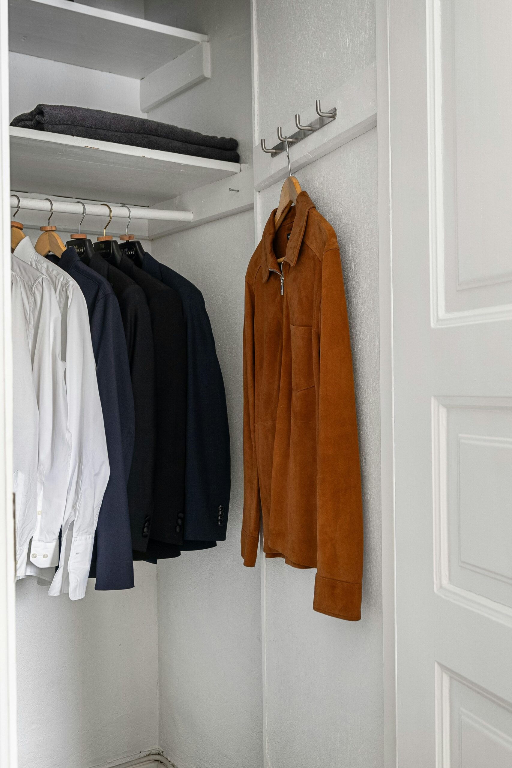 A small reach-in closet with white walls contains several white shirts, dark jackets on hangers, a brown suede jacket, and a folded dark item on the top shelf.