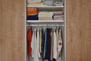 An open reach-in closet with folded towels and sheets on upper shelves and assorted shirts, jackets, and blouses hanging below.