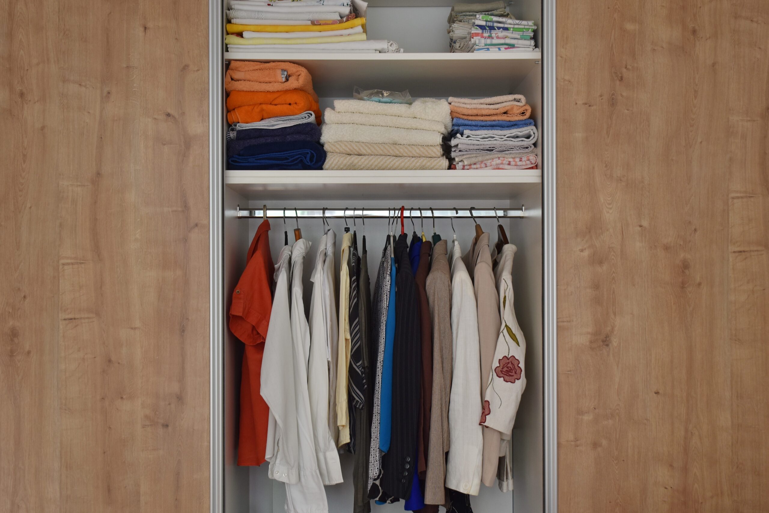 An open reach-in closet with folded towels and sheets on upper shelves and assorted shirts, jackets, and blouses hanging below.