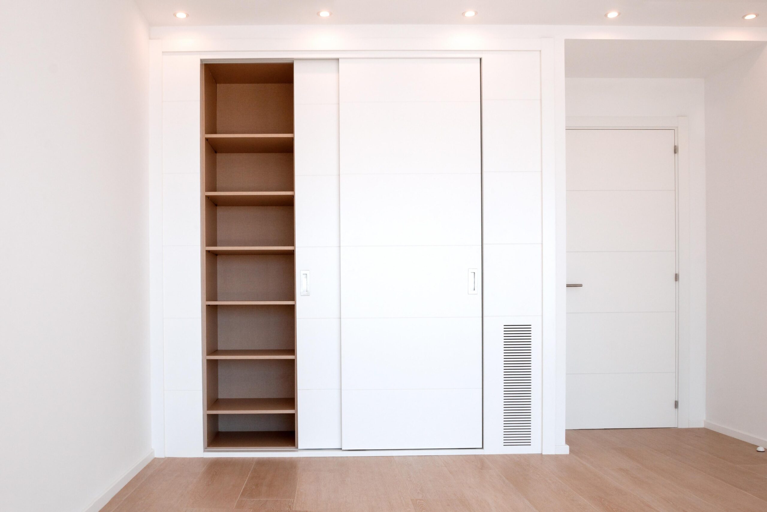 Minimalist white reach-in closet with sliding doors, revealing open shelves on the left; sits next to a closed white door in a bright, empty room with light wood flooring.