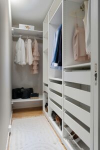 A neatly organized white walk-in closet, designed with reach-in closet functionality, features hanging clothes, handbags on a shelf, drawers, and a white rug on the floor.