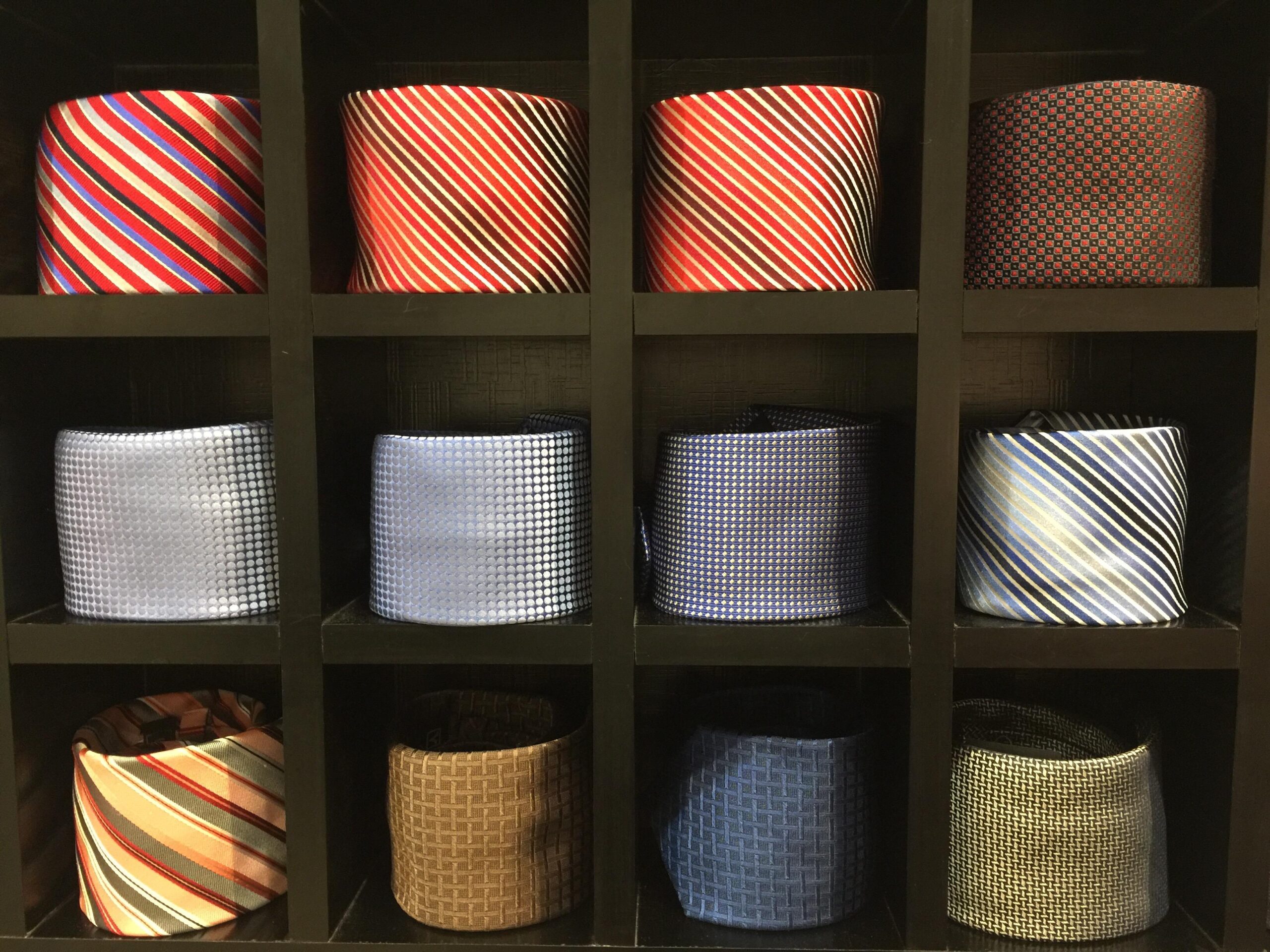 A display of twelve neatly rolled neckties in various colors and patterns arranged in a black grid shelf, perfect for organizing accessories in a reach-in closet.