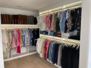 A neatly organized reach-in closet with clothes arranged by type and color on hangers, shelves with folded jeans, and tall brown boots lined up on the top shelf.