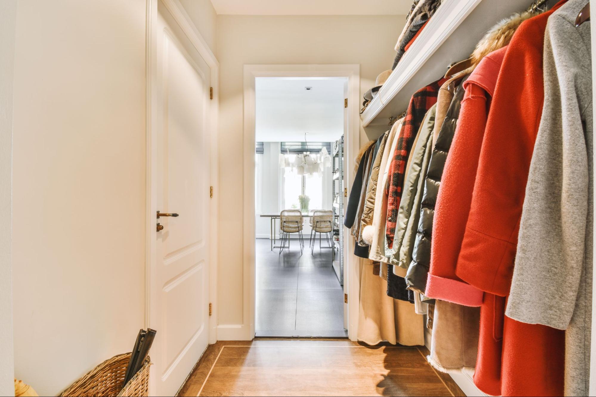 A hallway with a closed white door on the left and a rack of hanging coats on the right, leading to a bright dining area with a table and chairs.