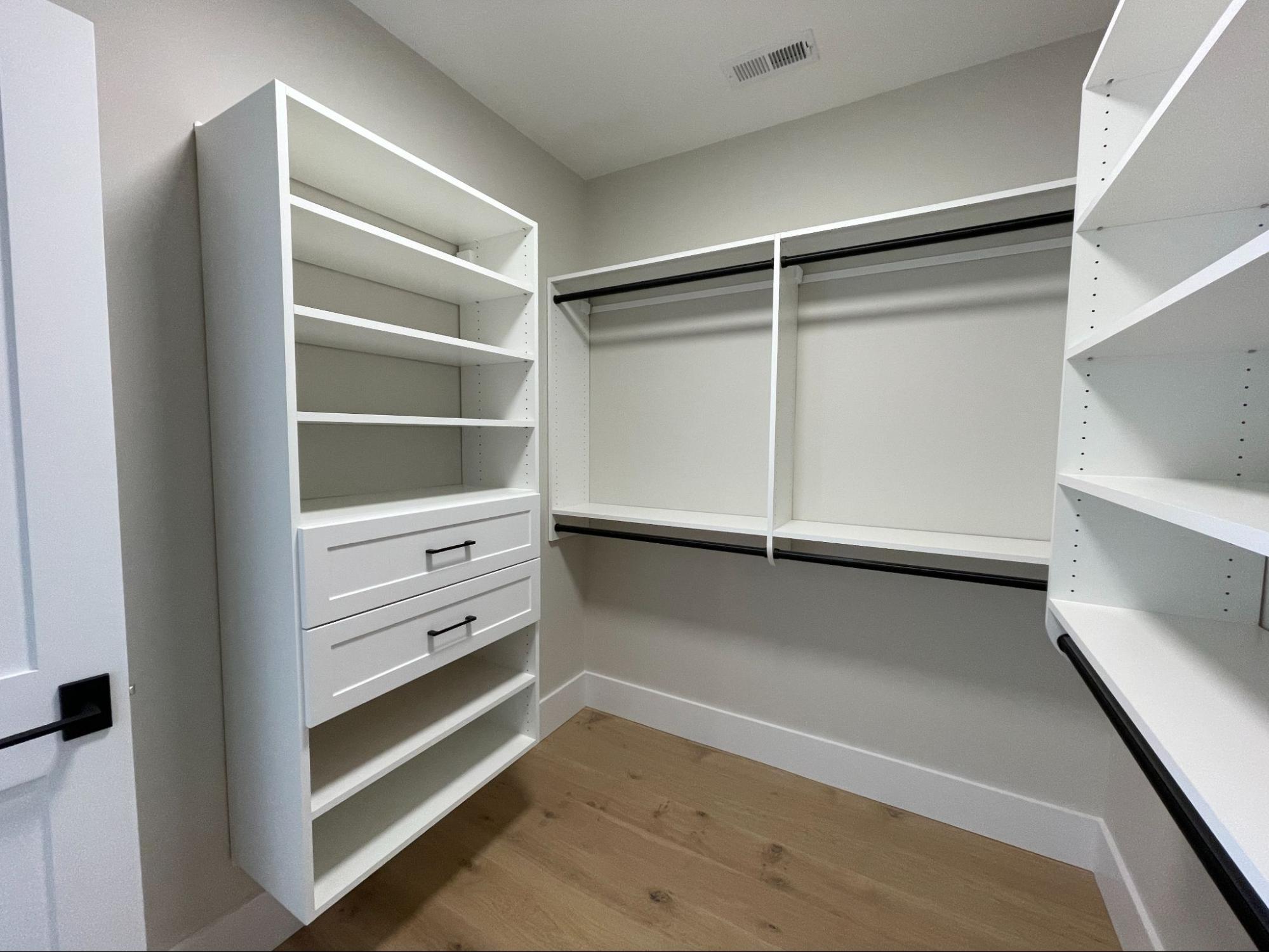 Empty walk-in closet with white built-in shelves, two drawers, hanging rods, and light wood flooring.