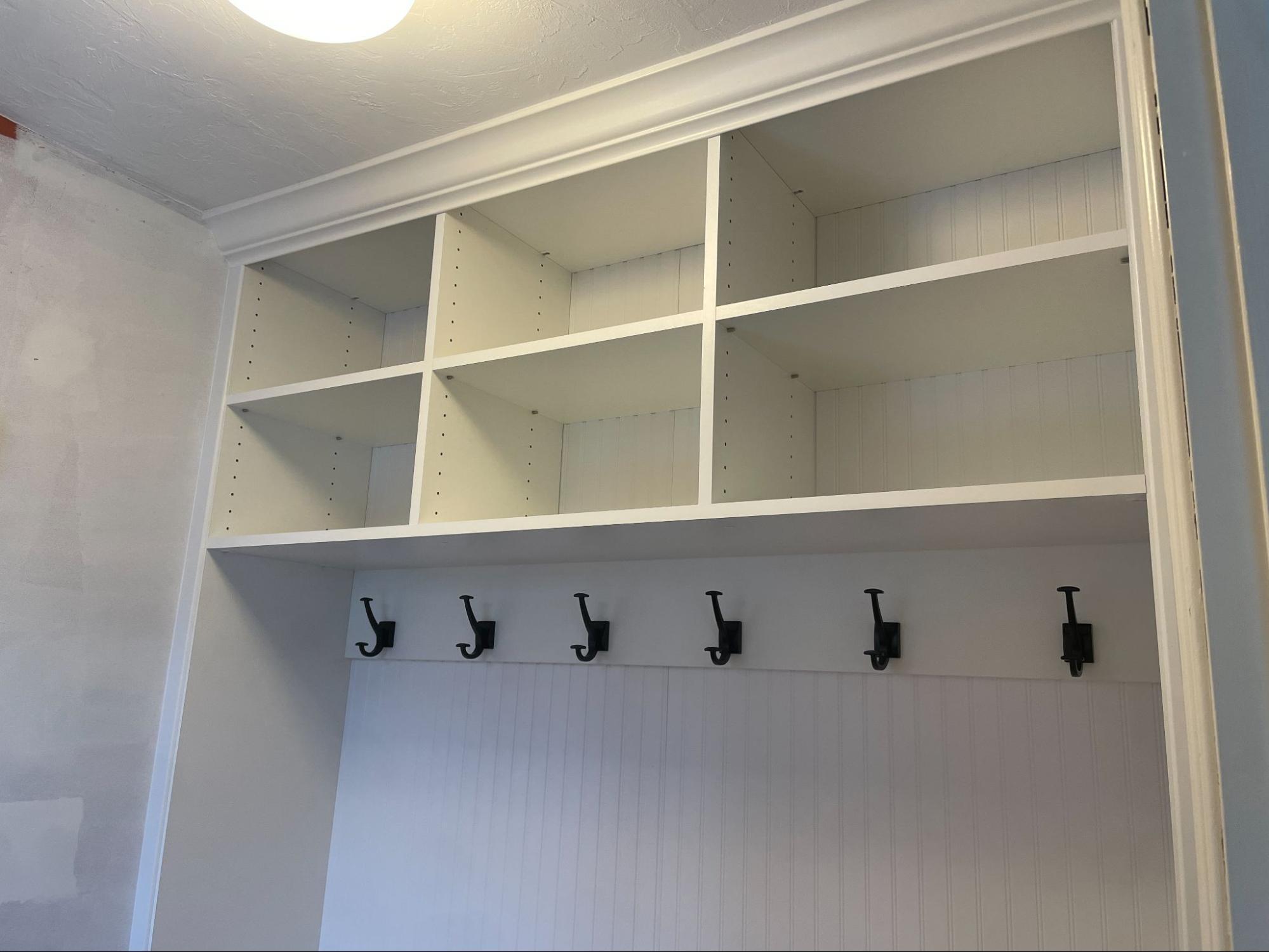 White built-in shelving unit with eight open cubbies above a row of six black hooks, against a white paneled wall in a room under renovation.