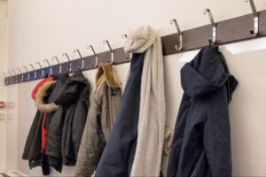Several winter coats, a scarf, and a knit hat hang on hooks mounted on a white wall.