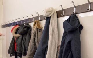 Several winter coats, a scarf, and a knit hat hang on hooks mounted on a white wall.