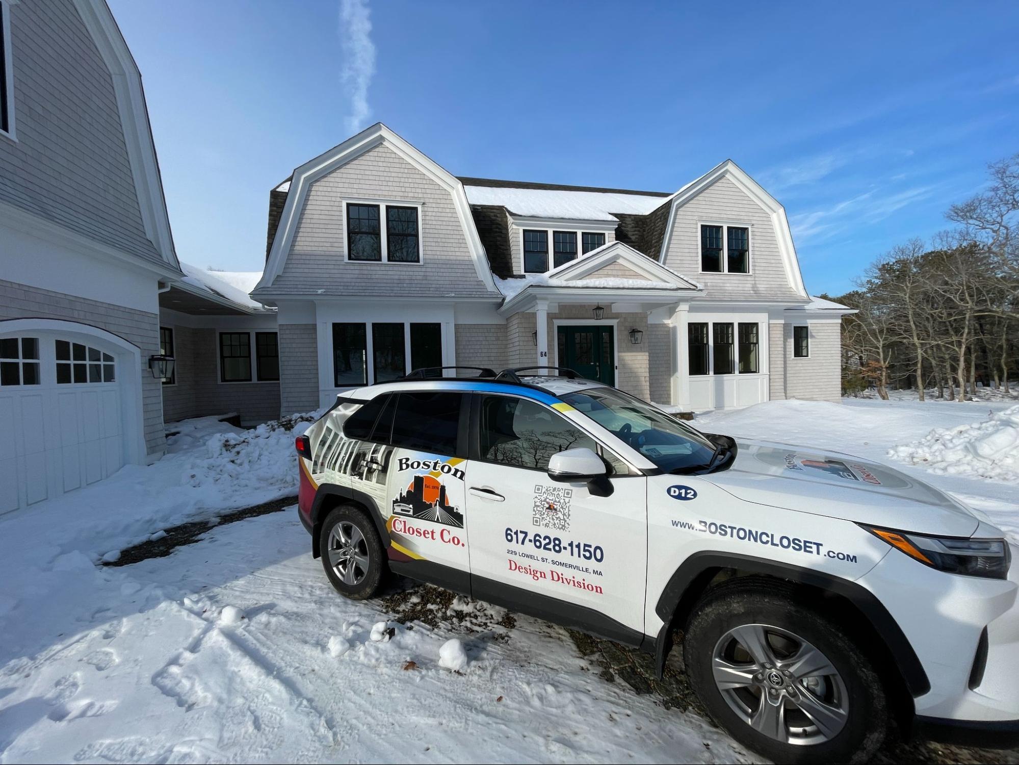 A Boston Closet Co. SUV is parked in front of a large, modern house with snow on the ground and a clear blue sky overhead.
