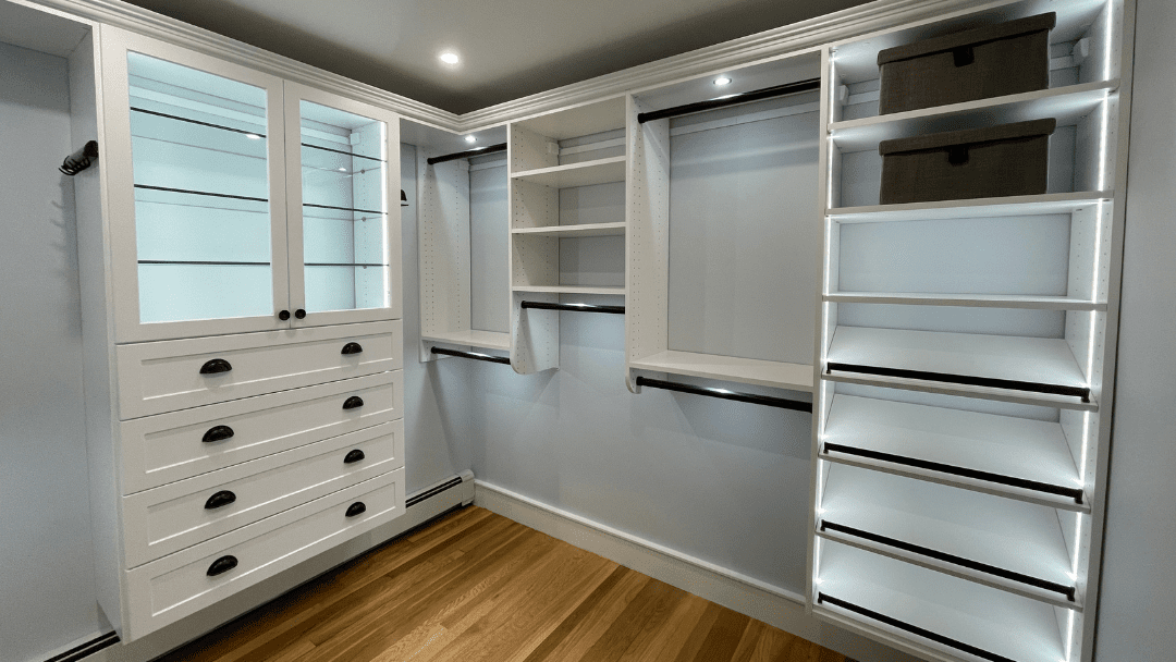 A modern walk-in closet with white shelves, drawers, hanging rods, and storage boxes, featuring built-in lighting and hardwood floor.