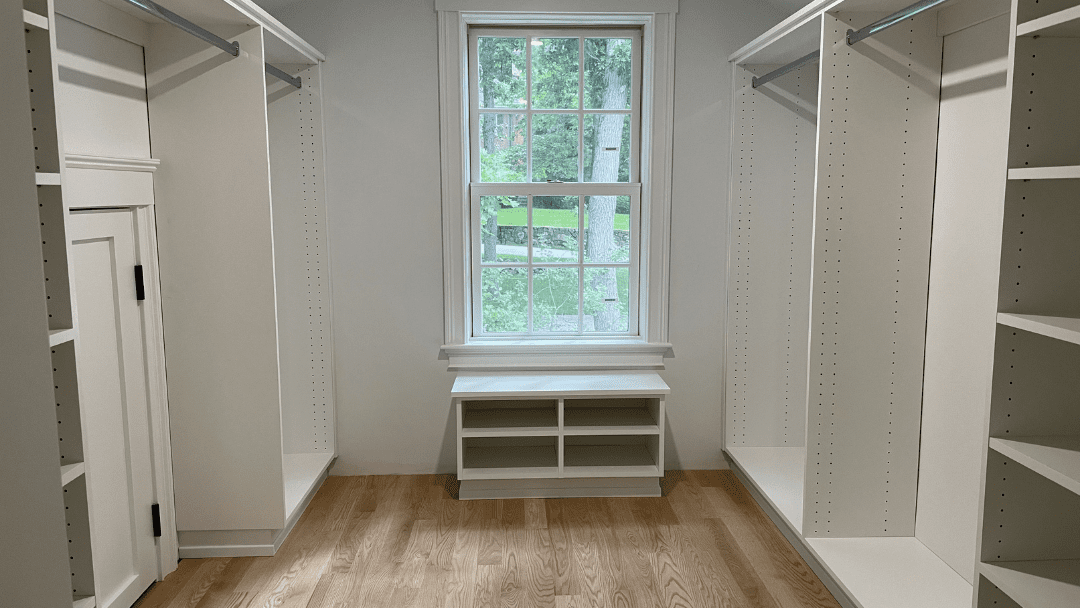 A walk-in closet with white shelves and rods, empty storage spaces, a window at the end, and light wooden flooring.